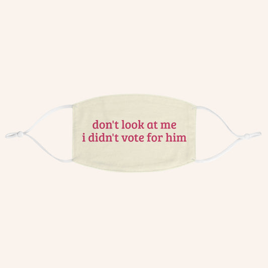 A cream-colored fabric face mask with white ear loops and pink printed text that reads 'don't look at me i didn't vote for him'.