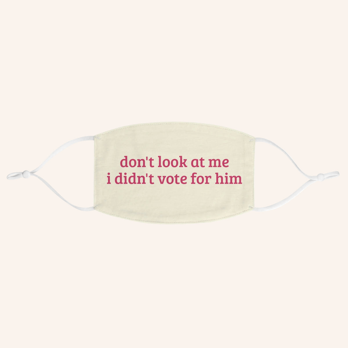 A cream-colored fabric face mask with white ear loops and pink printed text that reads 'don't look at me i didn't vote for him'.