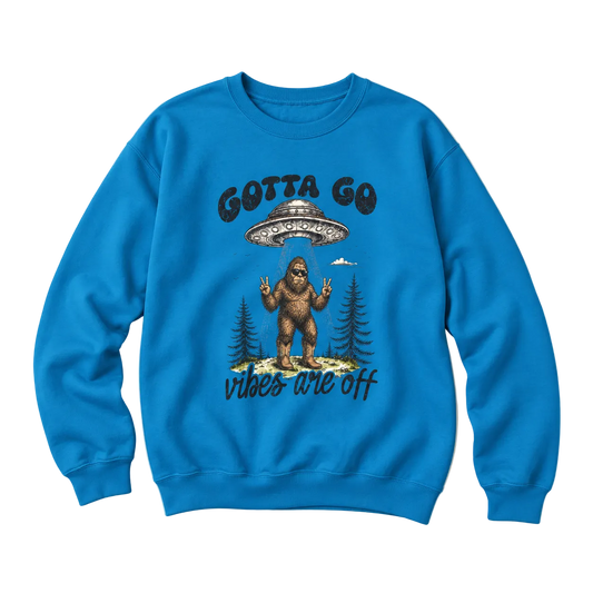 A bright blue crewneck sweatshirt featuring a graphic of Bigfoot under a UFO with the text 'GOTTA GO' above and 'vibes are off' below, displayed on a light cream background.