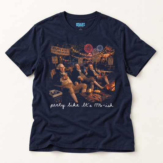 A navy blue short-sleeve graphic t-shirt featuring a vintage-style print of three men sitting by a campfire with 'Happy 4th of July' signs, and the text 'party like it's 1976-ish' printed below. The shirt is laid flat on a light beige background.
