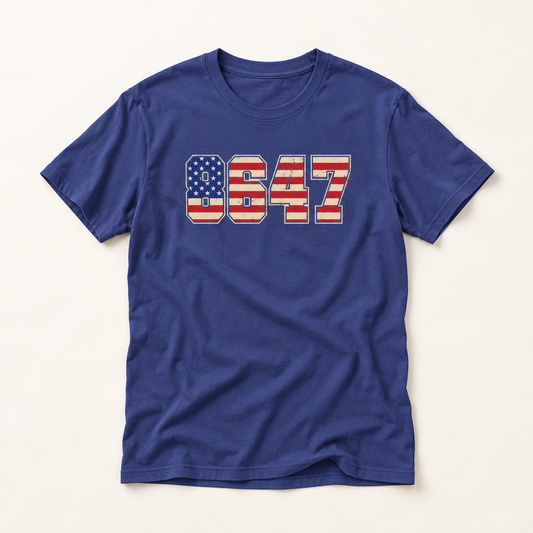 A royal blue short-sleeve t-shirt with a distressed graphic on the chest reading 'USA 8647' filled with an American flag pattern, displayed flat against a light beige background.