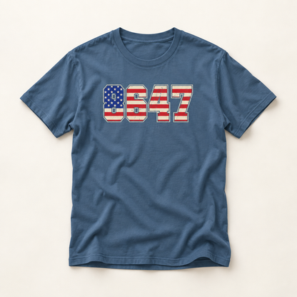 A blue crewneck t-shirt with a distressed graphic of the numbers '8647' filled with an American flag pattern, displayed flat against a light beige background.